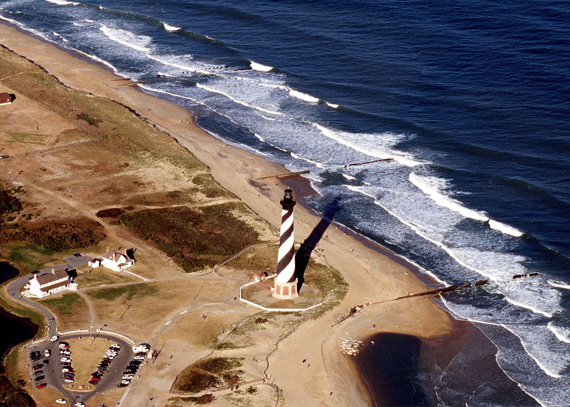 Aerial view of the black-and-white striped Cape Hatteras Lighthouse standing on a sandy beach next to the ocean.