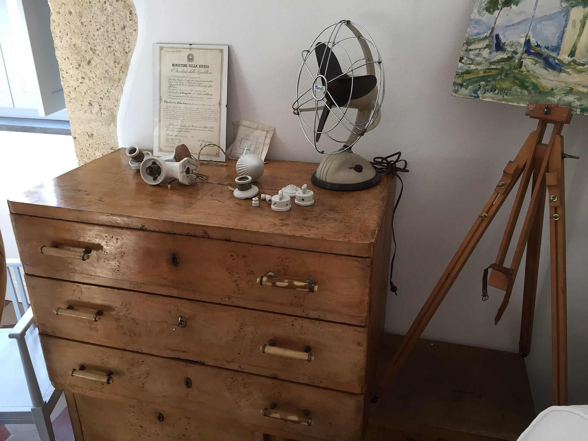 A wooden dresser holds a vintage electric fan, several ceramic electrical components, and a framed document, with an artist’s easel standing to the right.