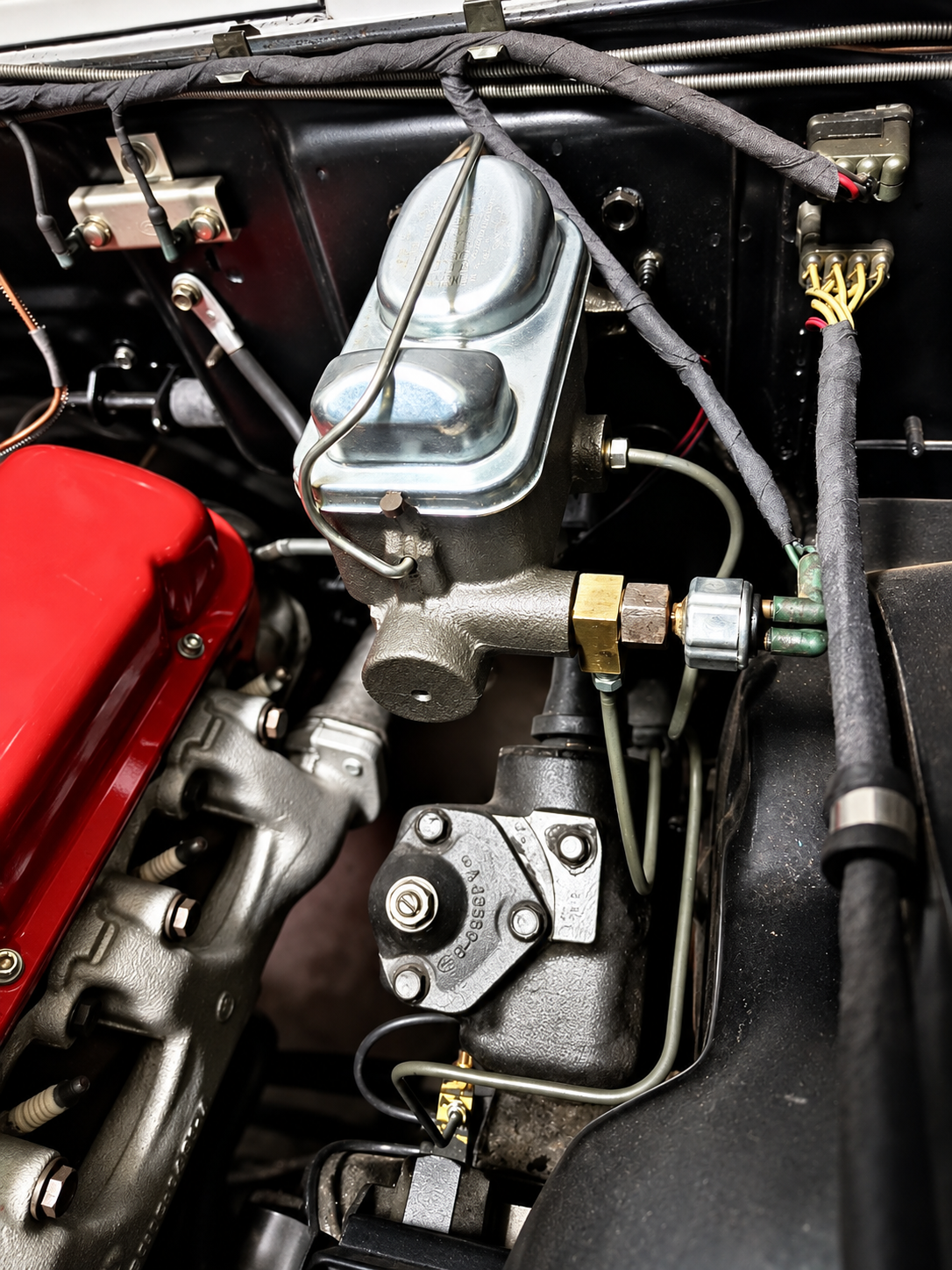 A close-up view of a car engine bay featuring a metal brake master cylinder with connected fluid lines, positioned next to a bright red valve cover.