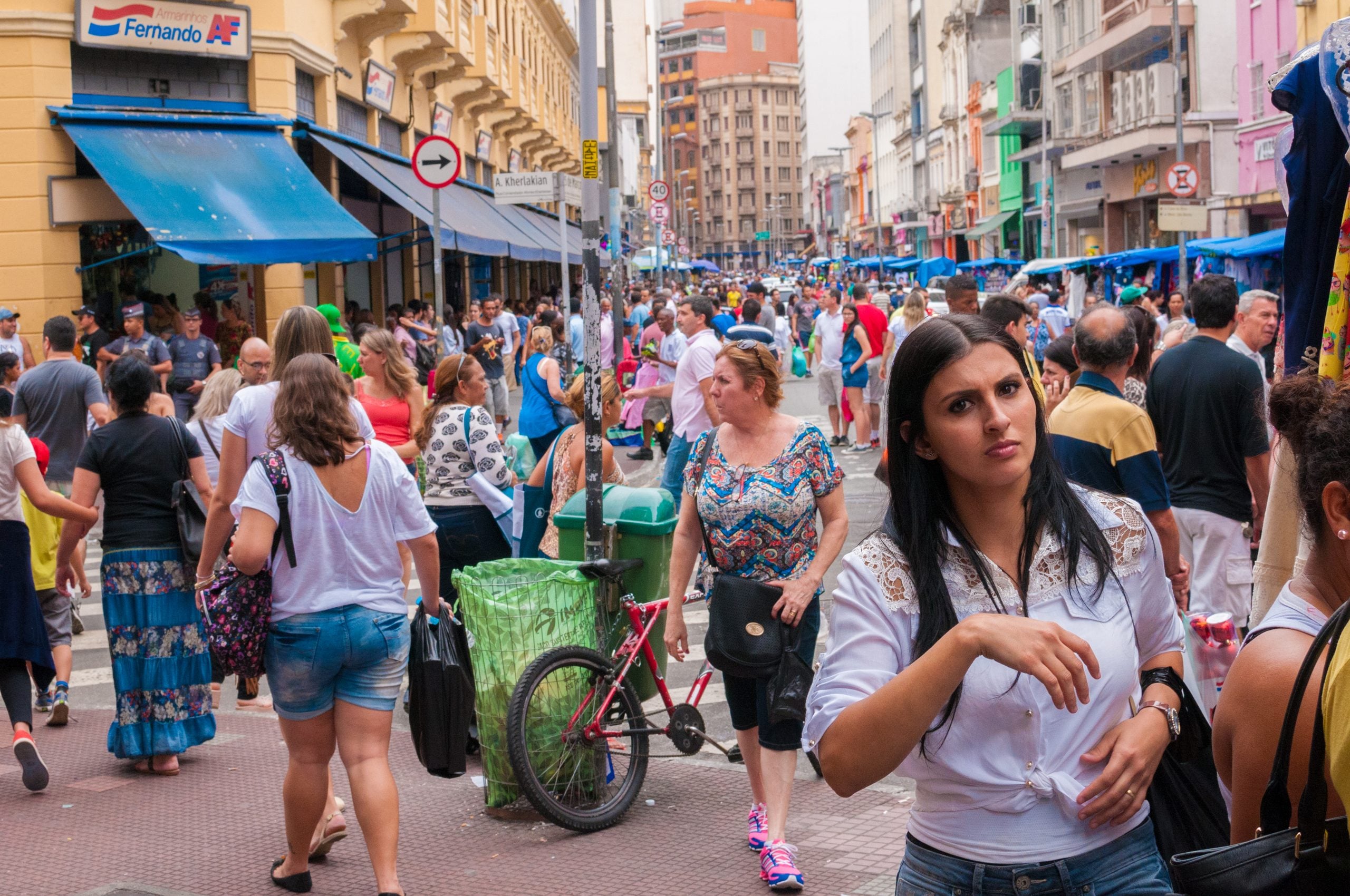 Crowded city street scene with many pedestrians walking along sidewalks lined with shops and blue awnings.