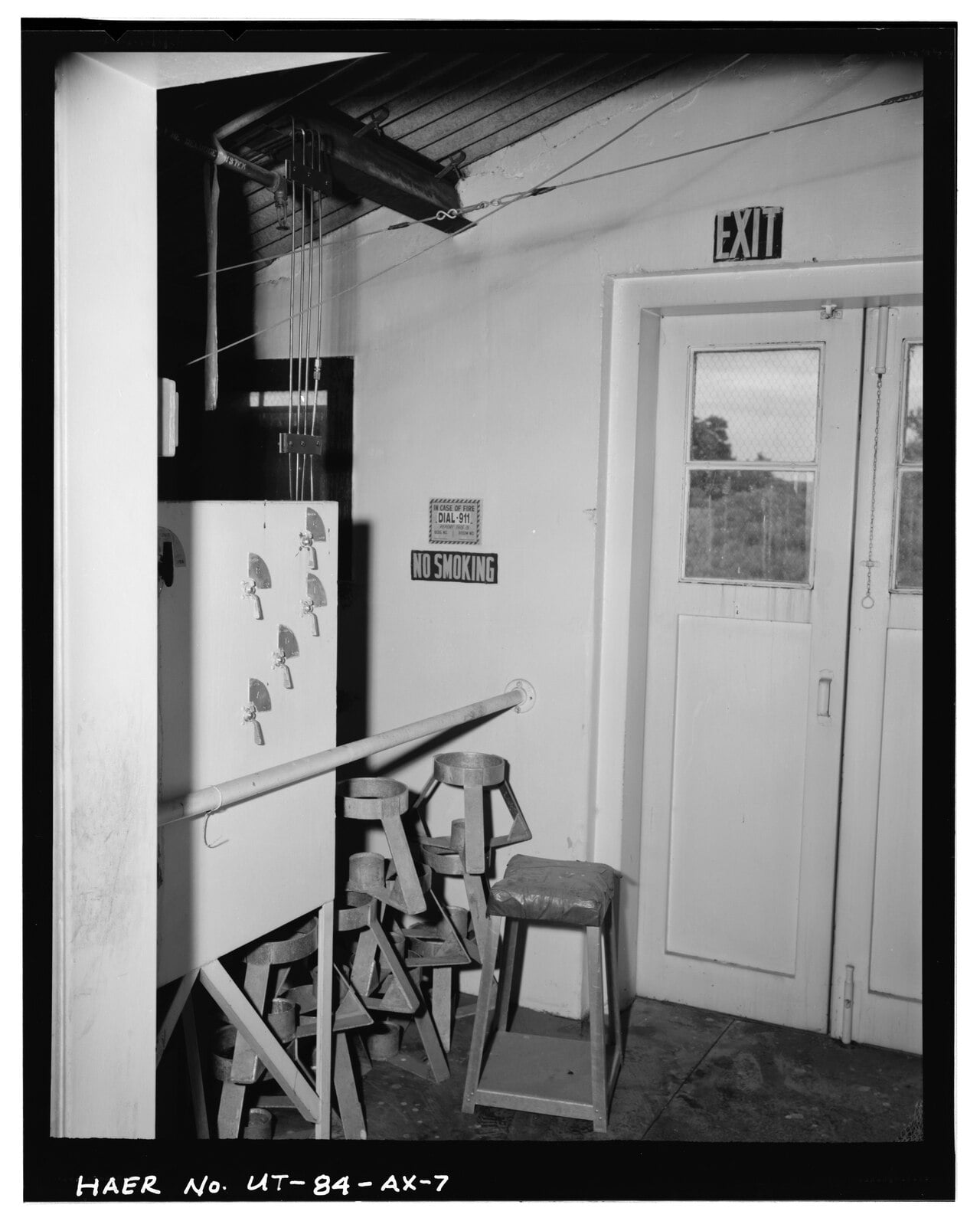 Industrial room interior featuring a wall-mounted panel with toggle switches, a handrail, several metal stands, a small stool, and a door with an “EXIT” sign above it.