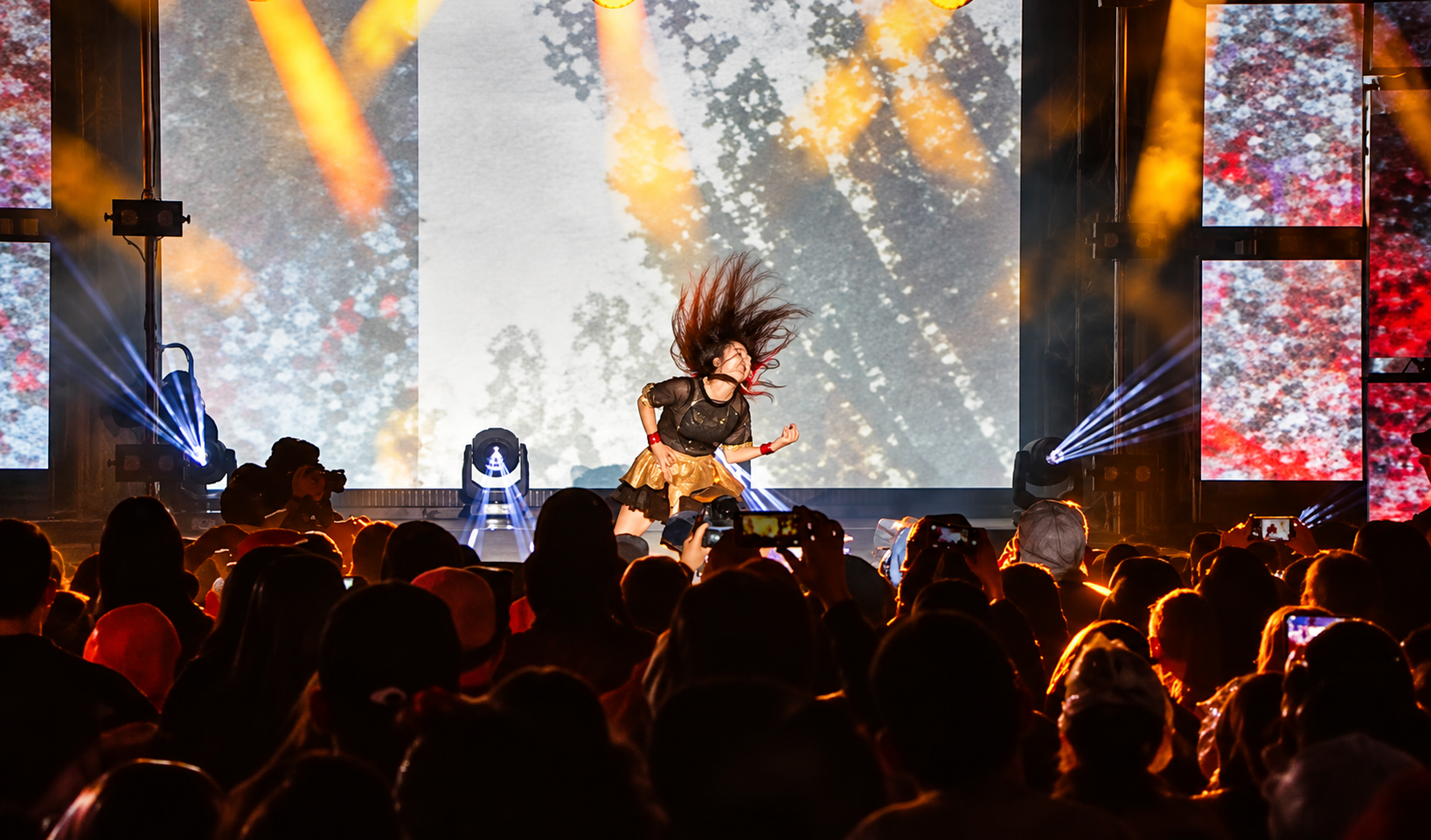 A performer on stage with hair mid-air, seen by a crowd holding up smartphones to record.