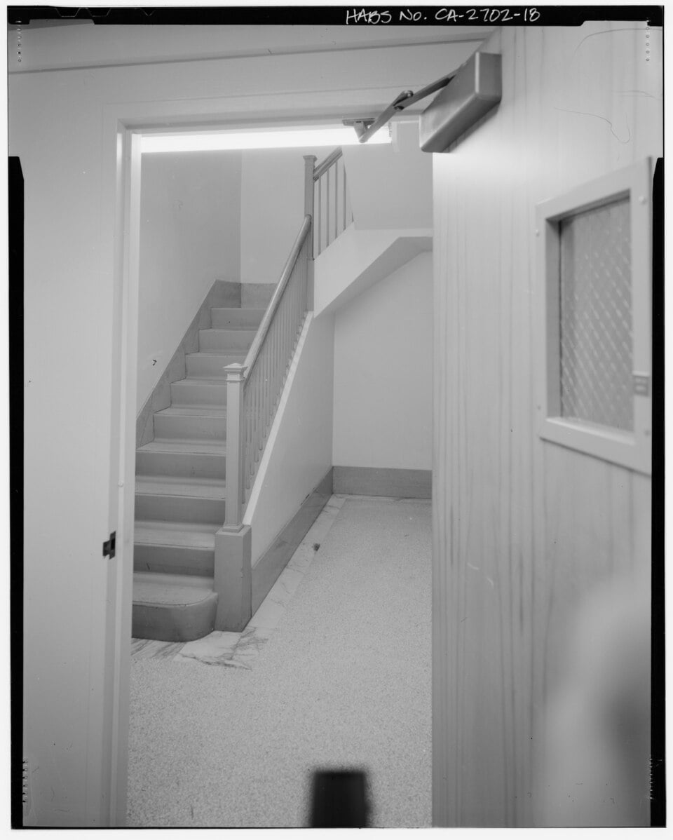 A view through an open doorway showing a stairwell with a wooden railing and a small window in the foreground door.
