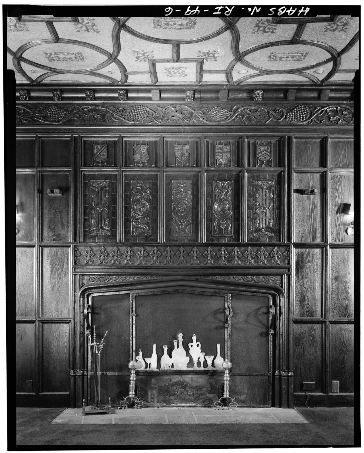 An ornate fireplace with dark wood paneling, featuring carved reliefs of crests and geometric patterns, with a collection of glass bottles arranged on the hearth.