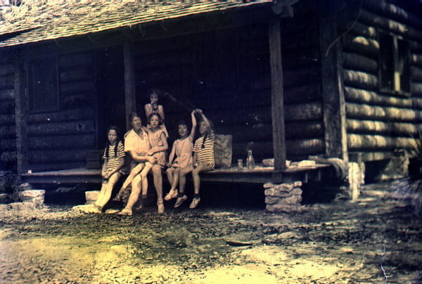 An adult woman sits on the porch of a log cabin surrounded by five young girls.