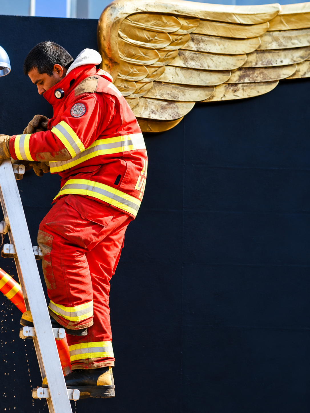 A firefighter in red protective gear climbing a metal ladder in front of a large gold wing sculpture on a black wall.