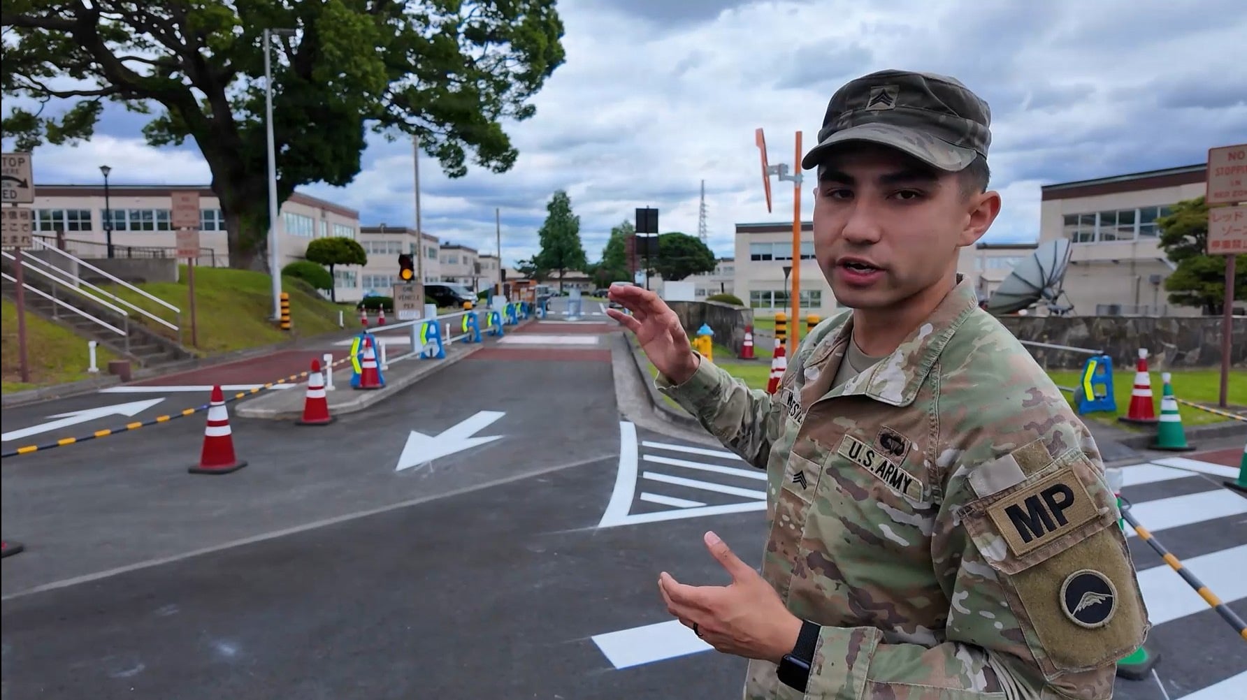A U.S. Army military police officer in camouflage uniform gestures toward a roadway with traffic cones and directional markings at a base entrance.