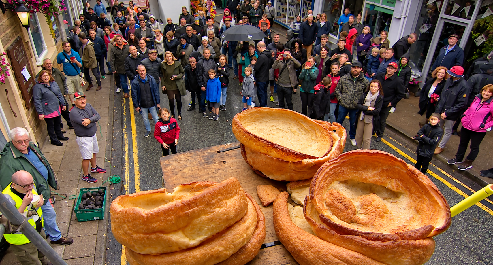 A large crowd gathers on a street to watch giant golden-brown Yorkshire puddings sitting on a wooden table.
