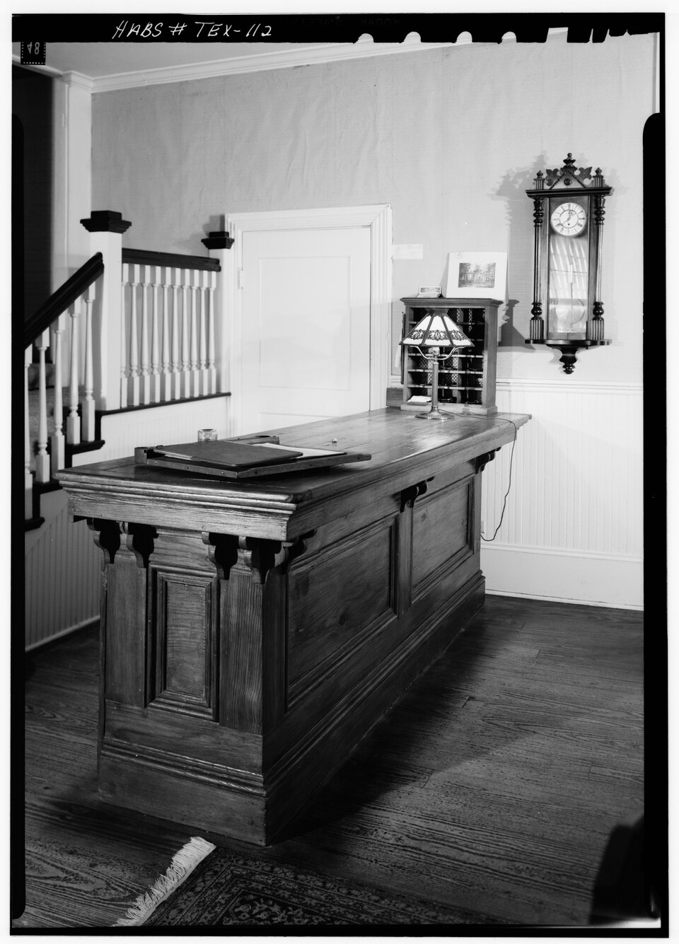 A long wooden reception desk sits in a hallway next to a staircase, with a small lamp, a pigeonhole mail organizer, and a wall clock nearby.