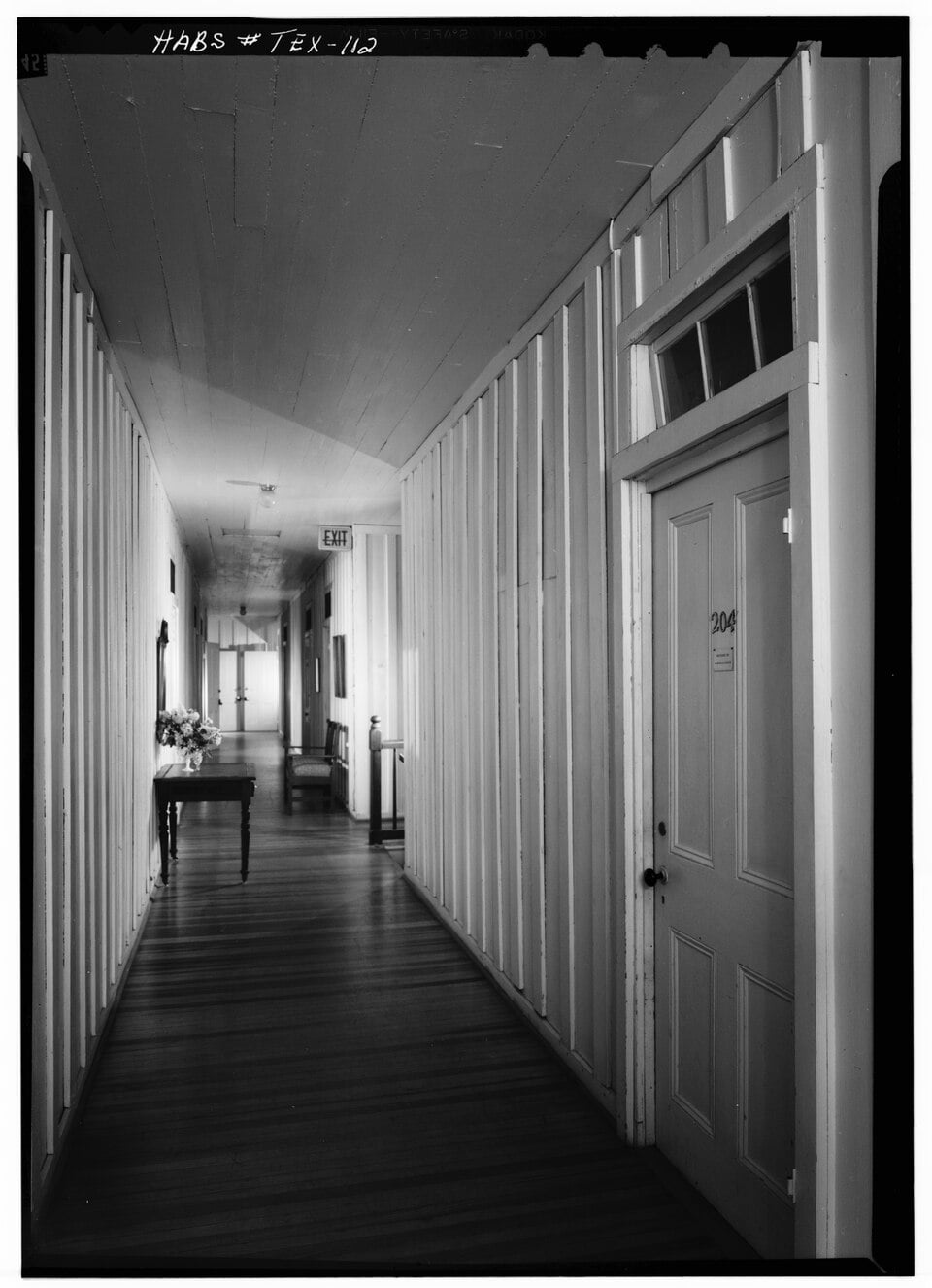 A long, narrow hallway with vertical wooden wall paneling, a wooden floor, and a door marked 204 on the right.