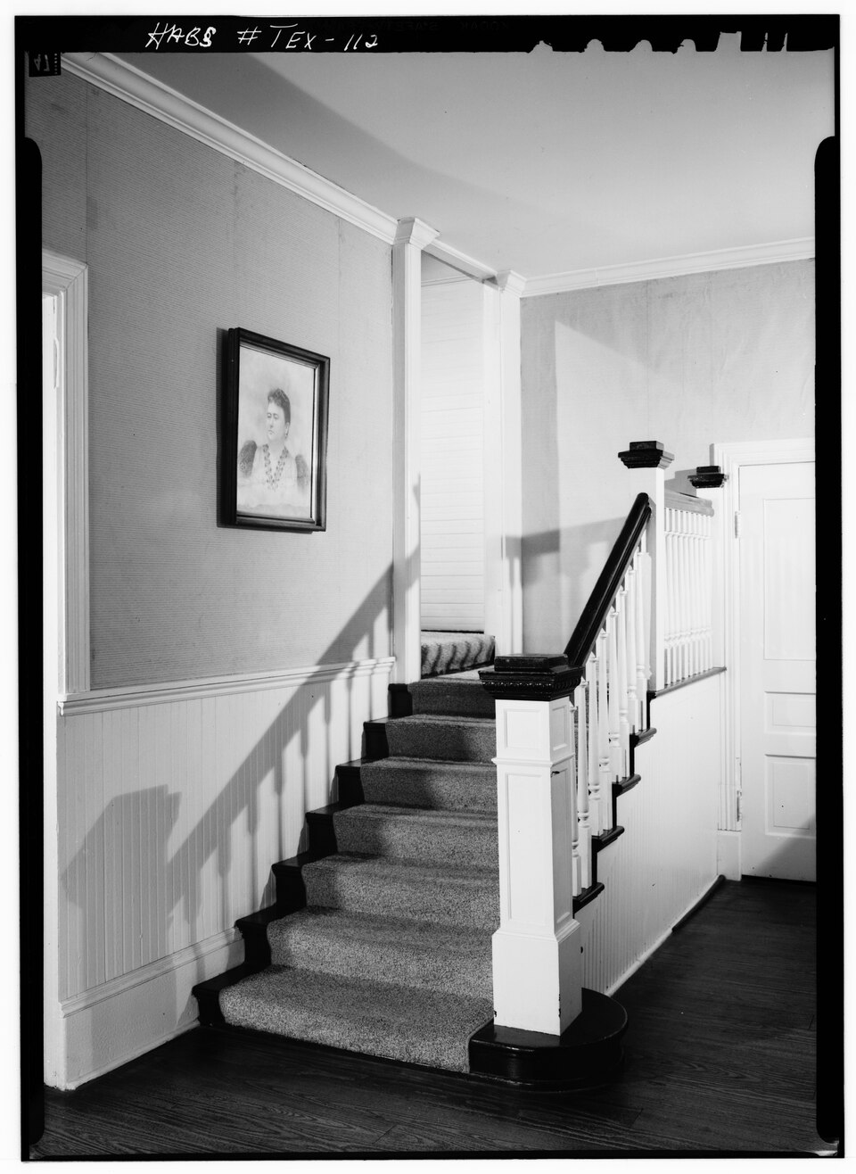 A staircase with a wooden handrail and carpeted steps, featuring a framed portrait on the wall and a doorway to the right.