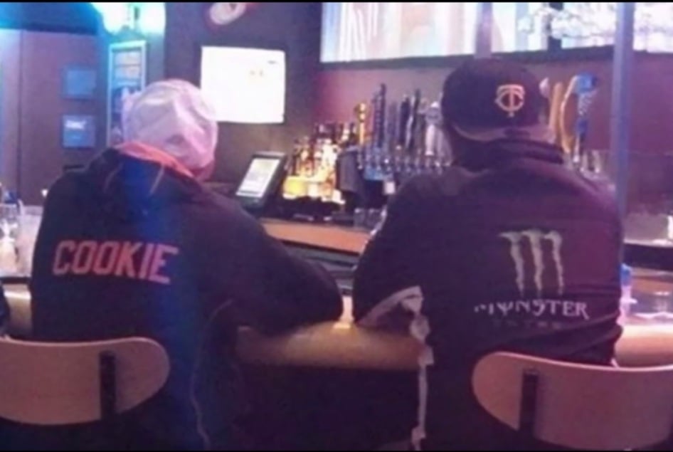 Two people seen from behind sit at a bar, one wearing a hoodie with the word “COOKIE” on the back and the other wearing a black Monster Energy hoodie and a baseball cap.