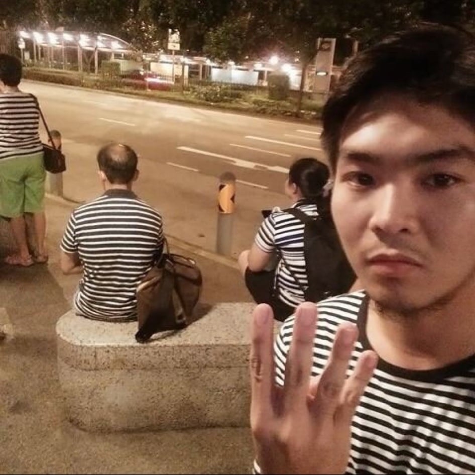 A selfie of a young man wearing a striped shirt and holding up four fingers, with people in matching striped clothing sitting on a stone bench behind him on a city street at night.