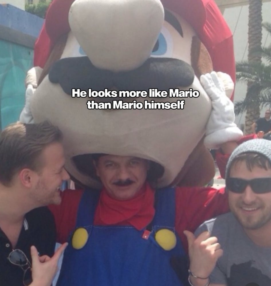 Two men pose with a person wearing a large Mario mascot head, with the text overlay: “He looks more like Mario than Mario himself.”
