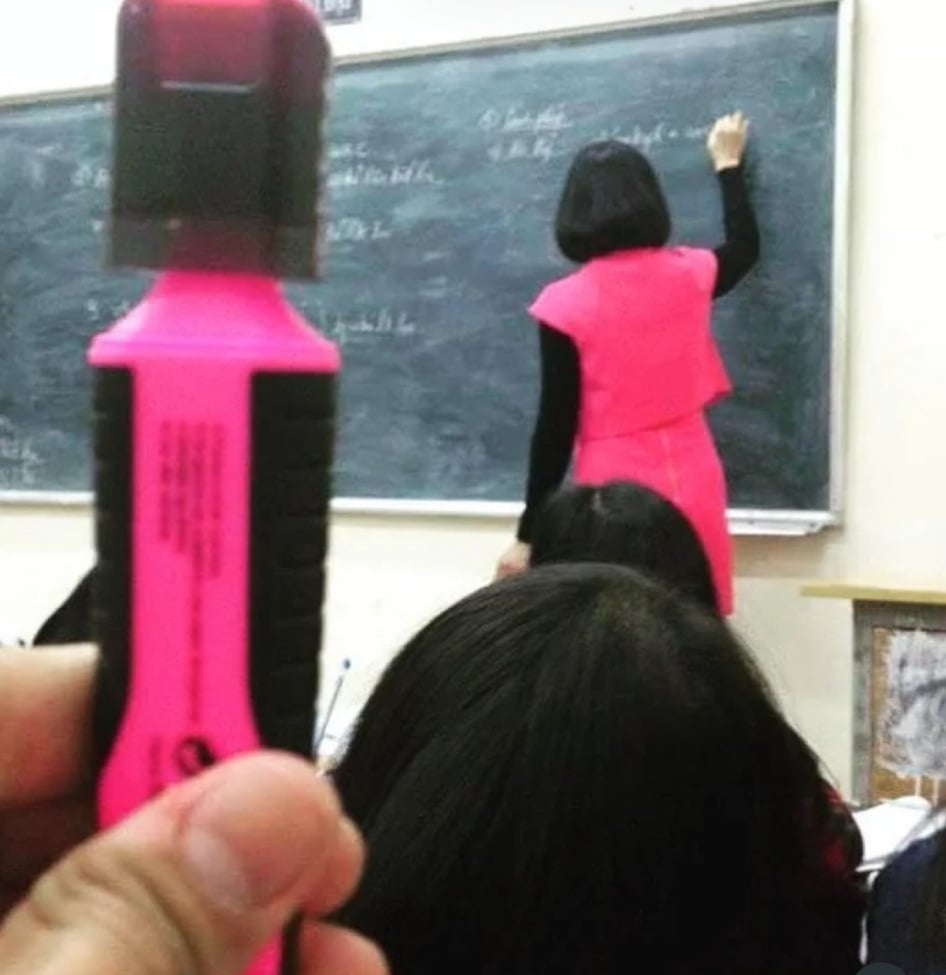 A person in the foreground holds a bright pink highlighter, while a teacher in a matching pink vest writes on a chalkboard in the background.