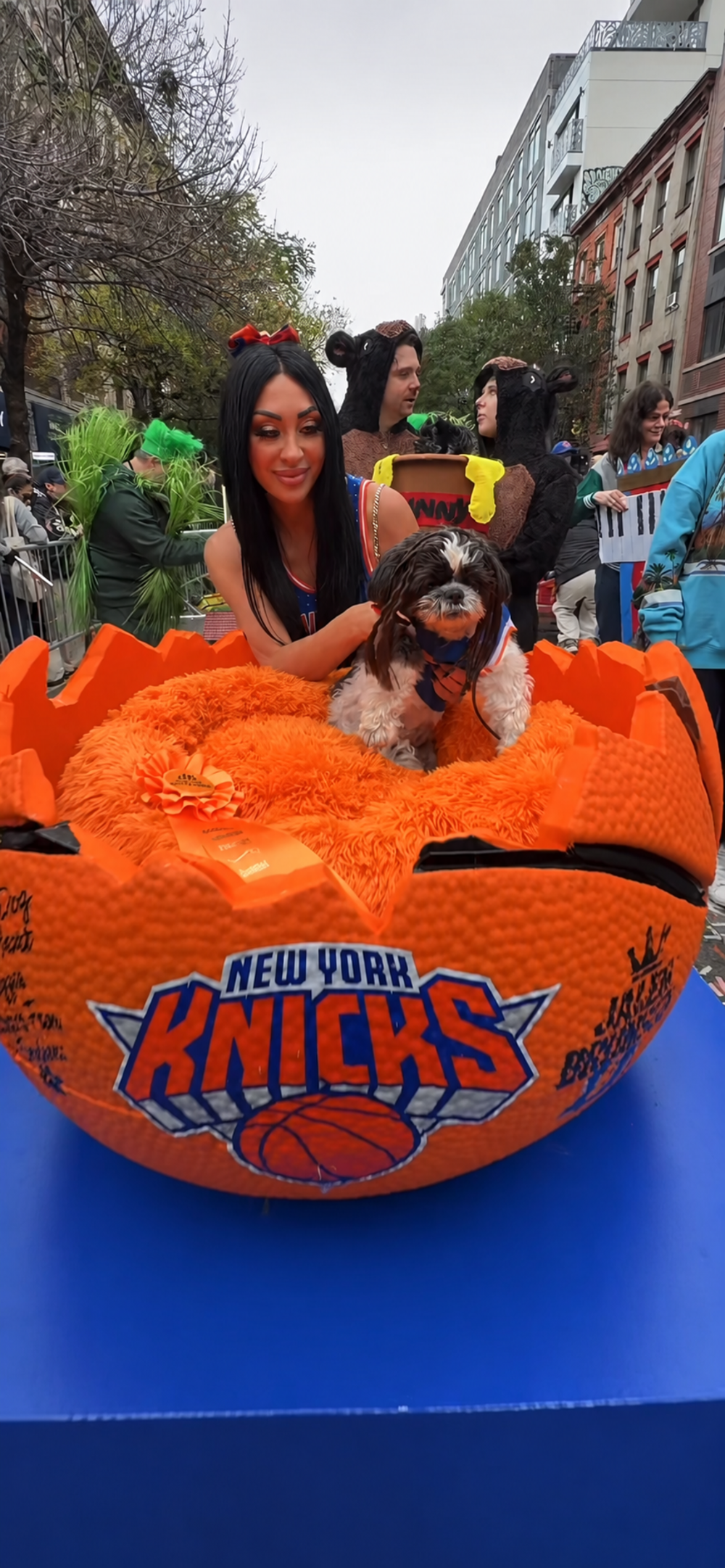 A woman in a New York Knicks jersey sits behind a large, orange basketball-shaped pet bed, petting a small dog that is sitting inside the bed.