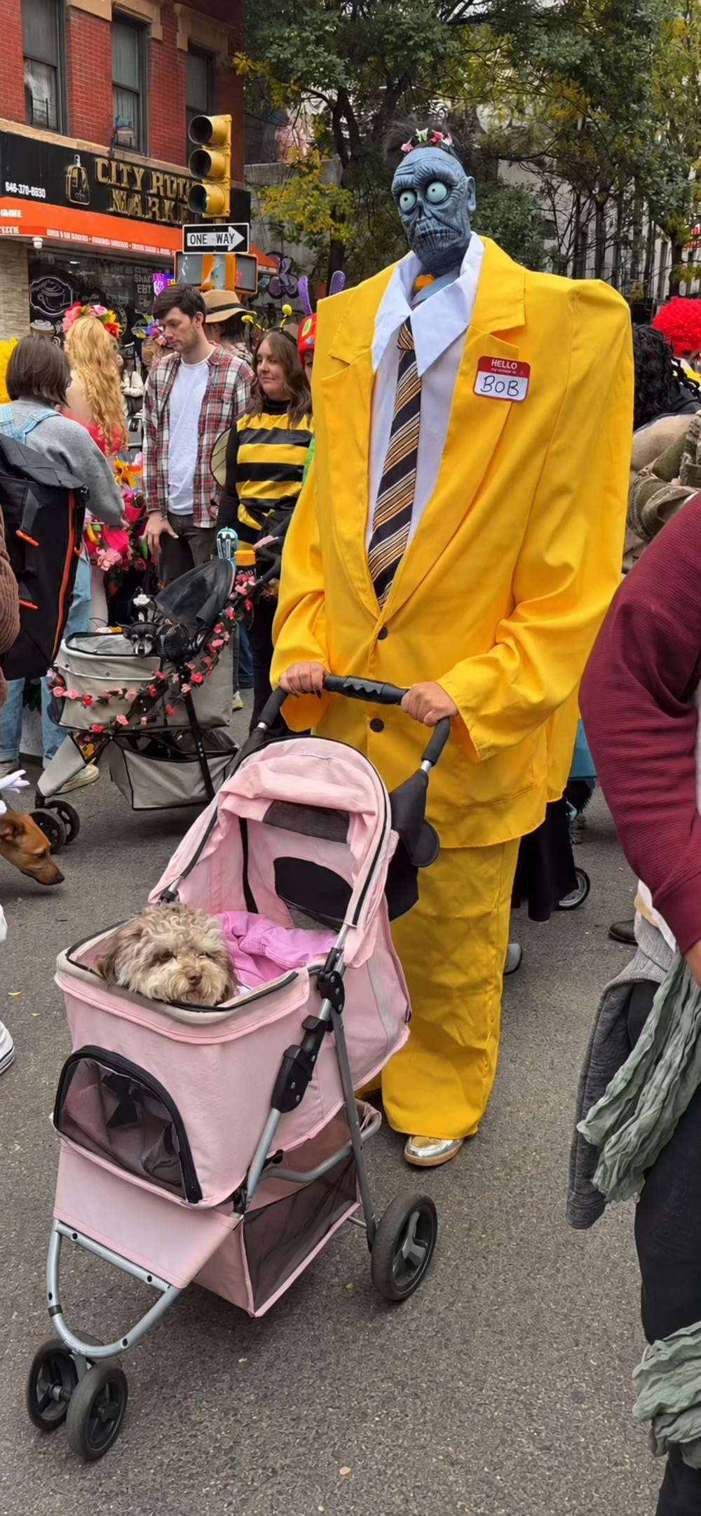 A person dressed as a zombie in an oversized yellow suit with a “Hello My Name Is Bob” nametag pushes a small dog in a pink pet stroller.