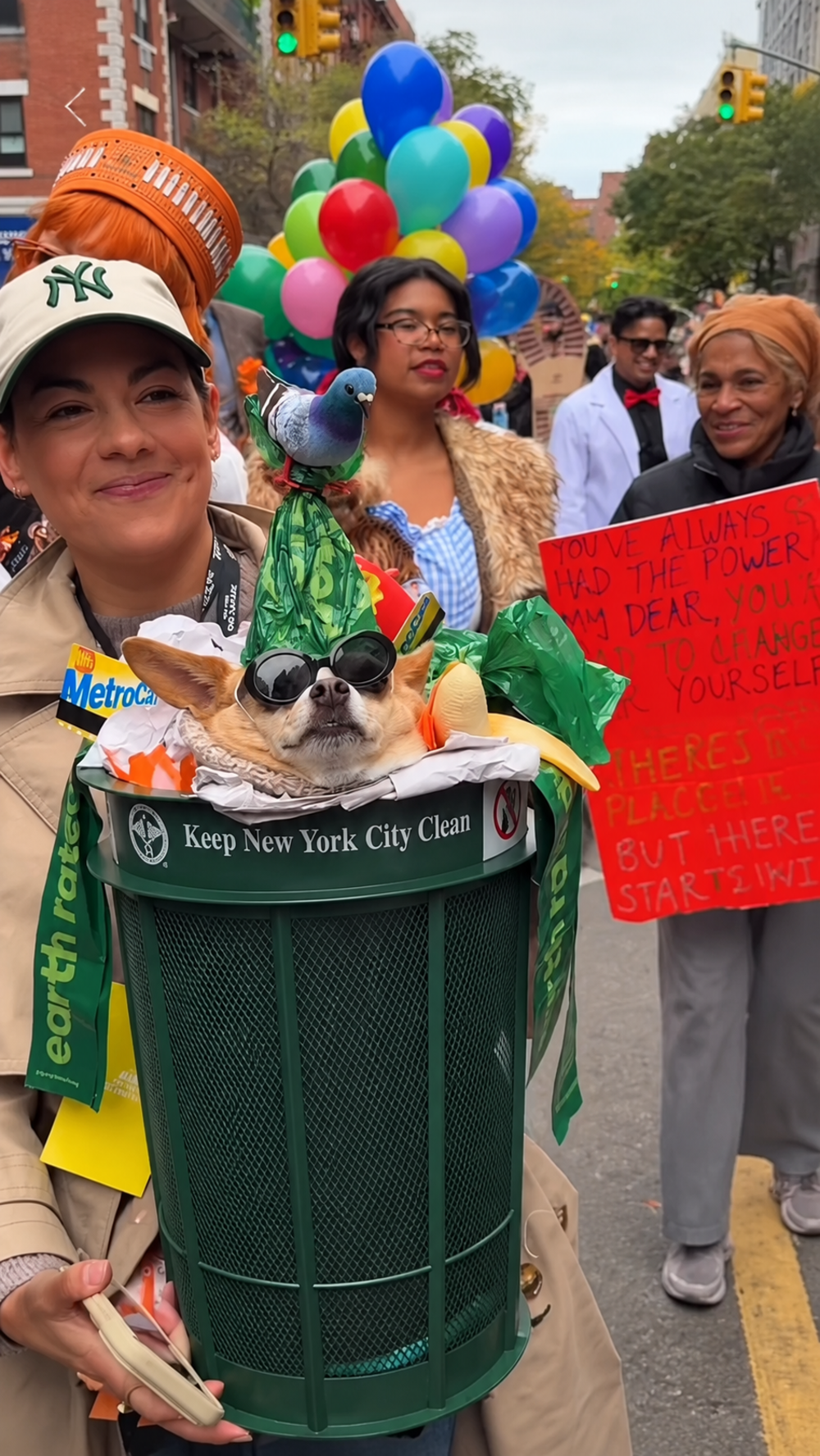 A woman in a baseball cap carries a chihuahua wearing sunglasses and a pigeon hat inside a green New York City trash can at a parade.