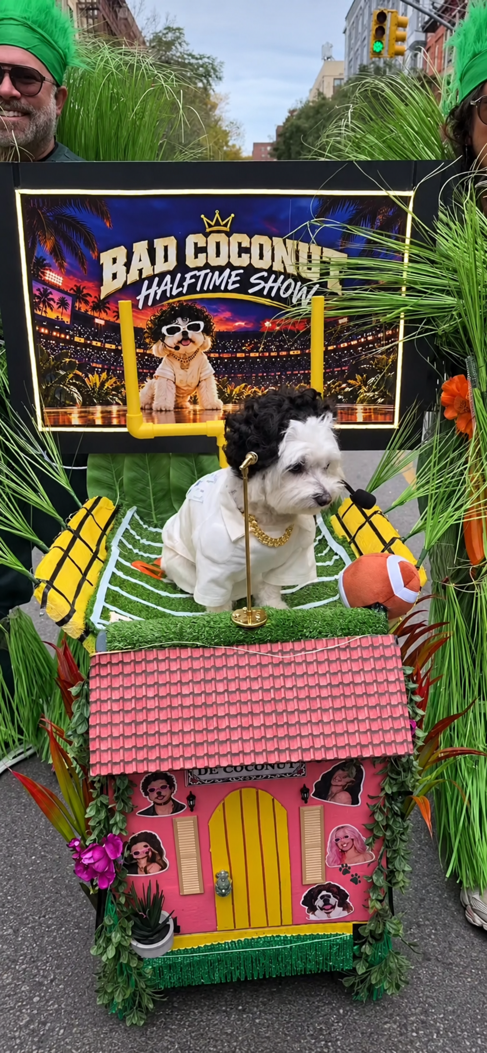 A small white dog wearing a black wig and gold chain sits on a platform shaped like a football field, in front of a “Bad Coconut Halftime Show” sign and a pink house prop.