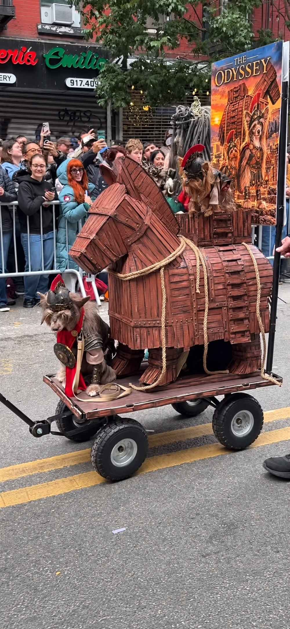Two dogs dressed as ancient warriors riding in a wooden Trojan horse replica on a wheeled cart during a parade.