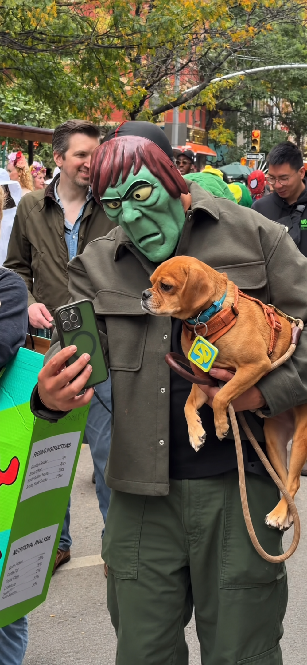 A person wearing a green monster mask holds a small tan dog while looking at a smartphone screen.