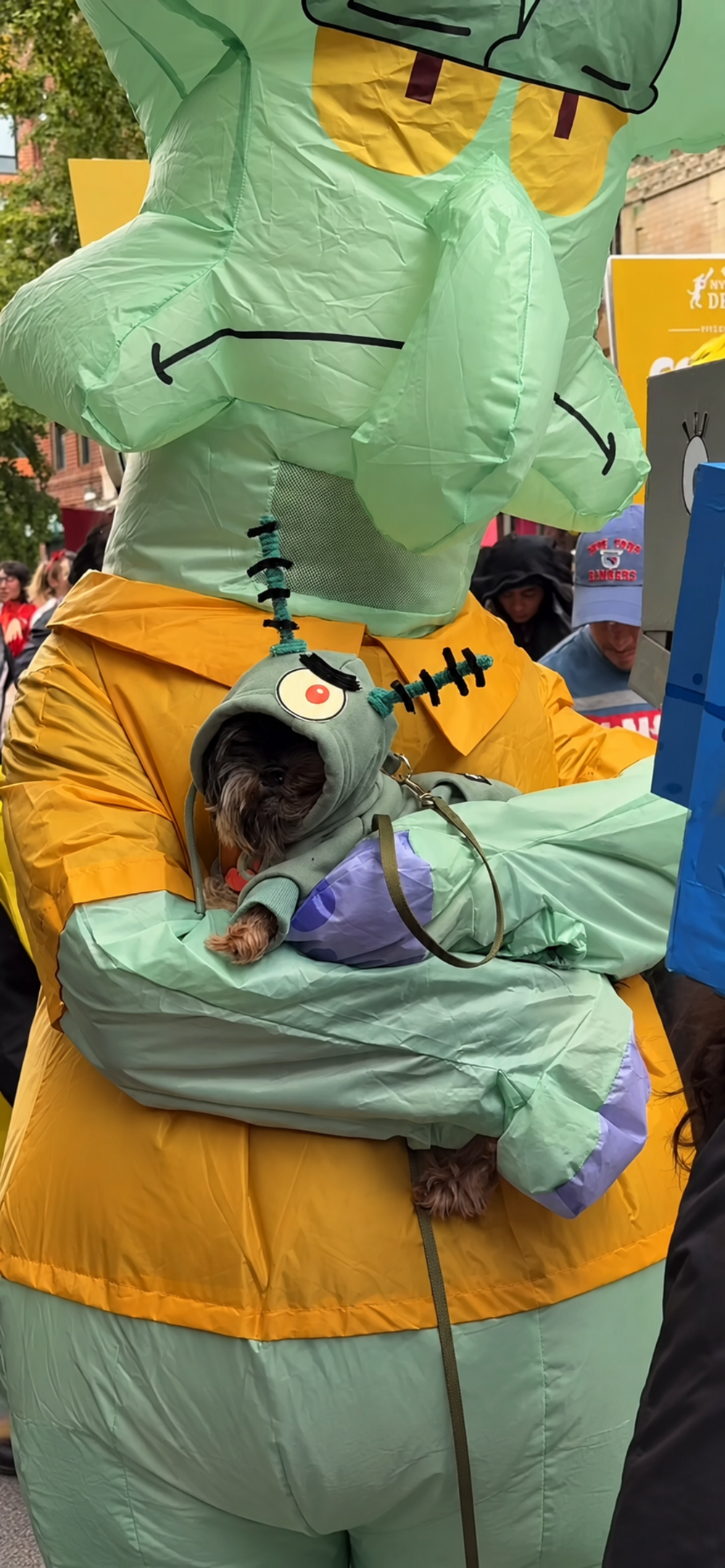 A person in an inflatable Squidward costume holds a small dog wearing a green Plankton-themed hoodie.