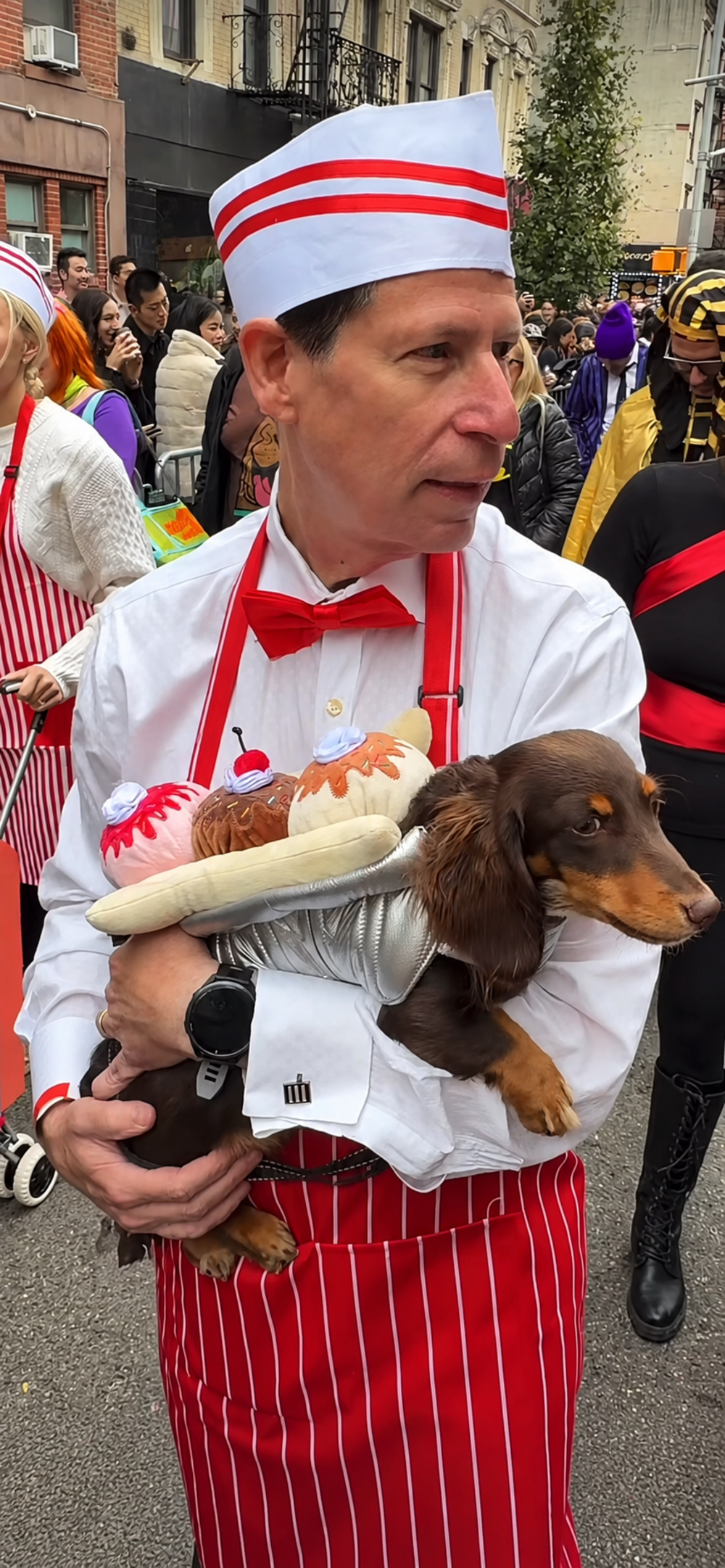 A person dressed as an ice cream parlor server holds a dachshund wearing a banana split costume.