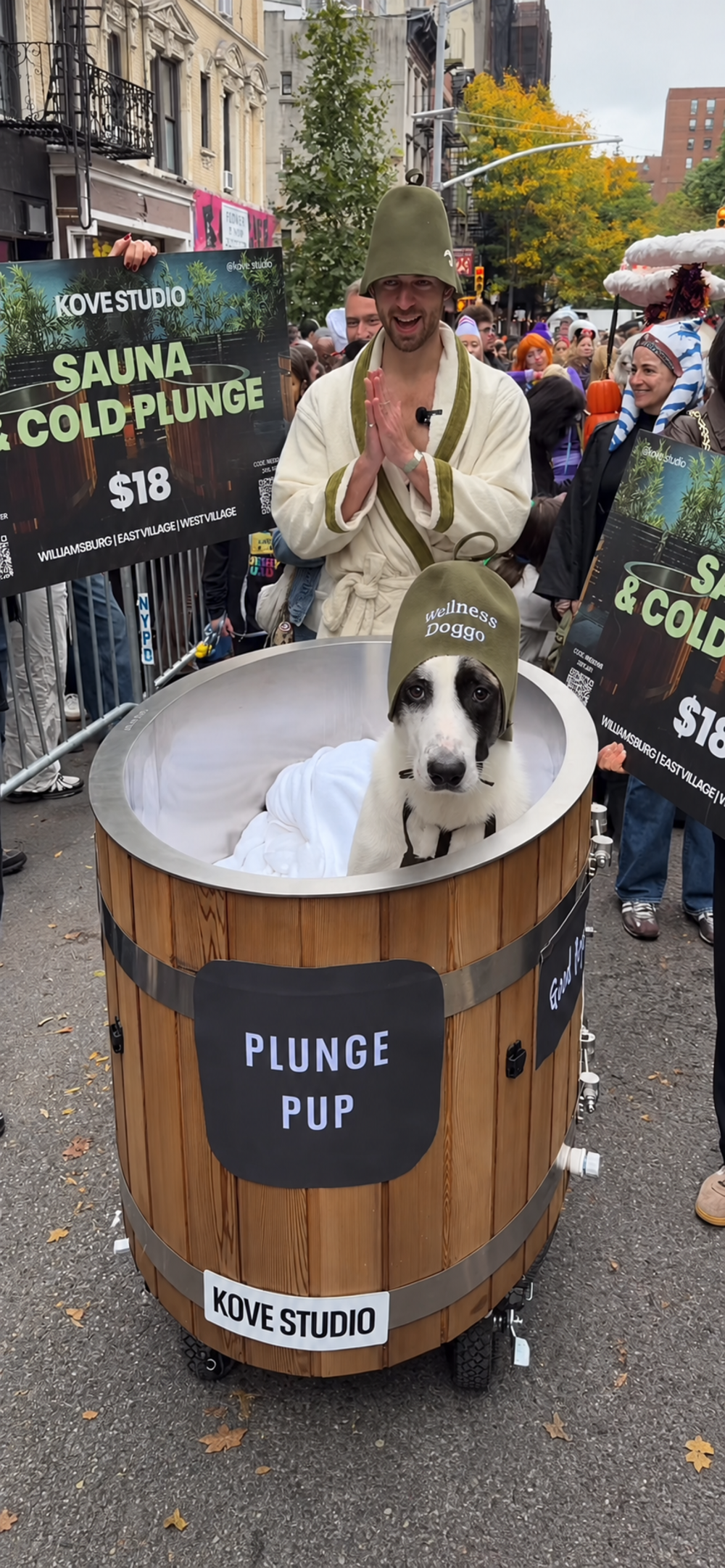 A dog wearing a branded beanie sits inside a portable wooden cold plunge barrel, with a man in a matching hat and bathrobe standing behind it.