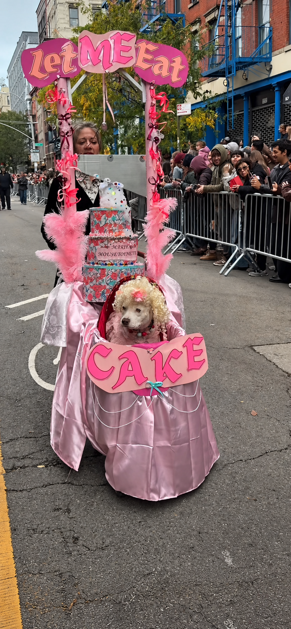 A dog dressed as Marie Antoinette sits in a pink, cake-shaped parade float labeled “LetMEEat” and “CAKE.”