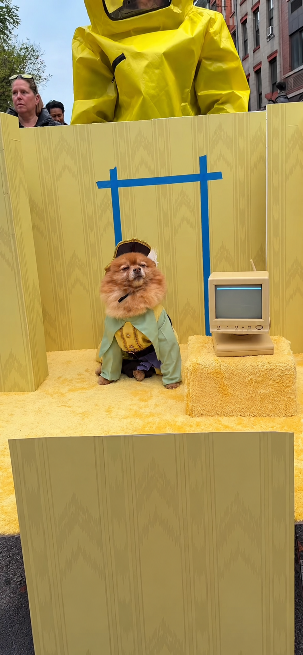A Pomeranian wearing a military-style hat and jacket sits on a yellow stage next to a vintage computer monitor, with a person in a yellow hazmat suit standing behind the set.