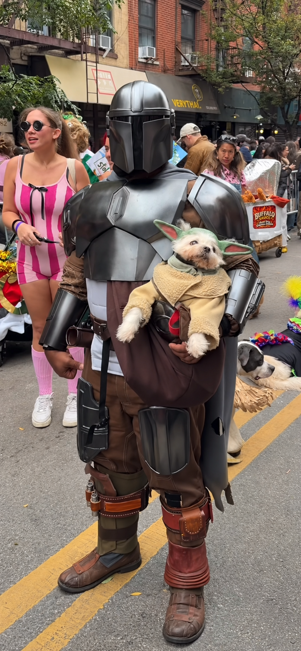 A person in a Mandalorian costume holding a small dog dressed as Grogu in a sling, standing on a city street.