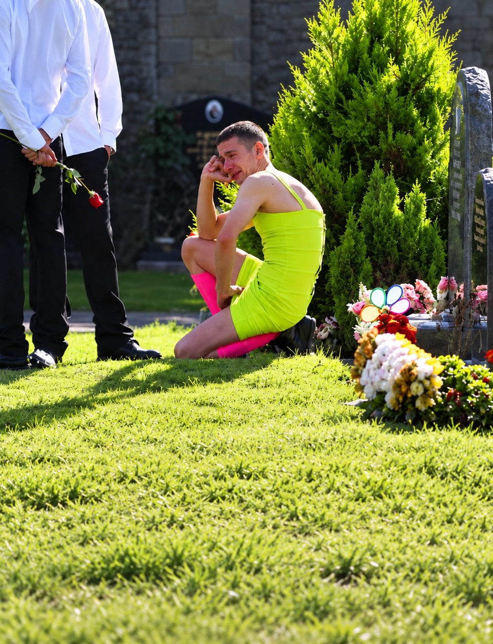 A man in a neon yellow dress and bright pink leg warmers kneels crying at a grave site, while a person standing next to him holds a red rose.