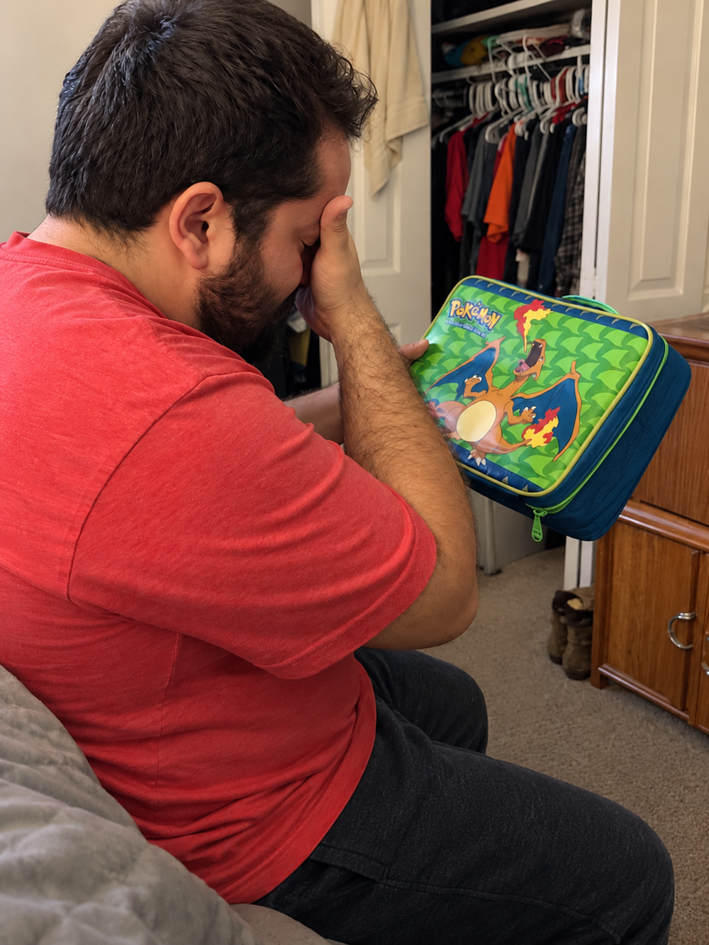 A man in a red shirt holds his head in his hand while looking at a Pokémon lunchbox featuring Charizard.