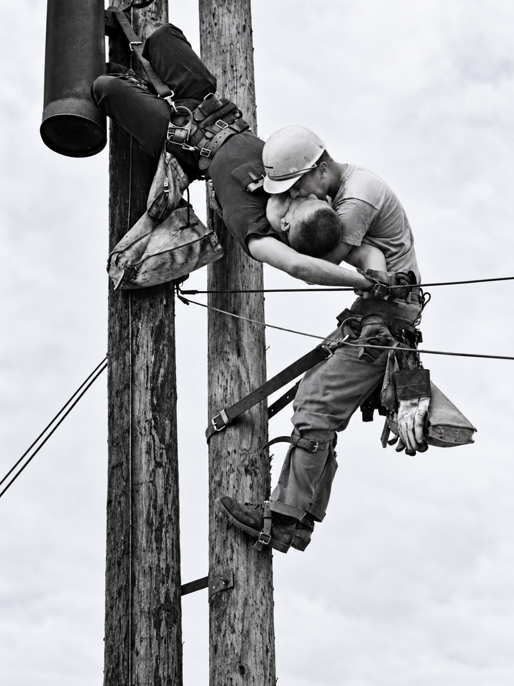 A utility worker suspended on a wooden pole kisses a fellow worker who is attached to the same pole.