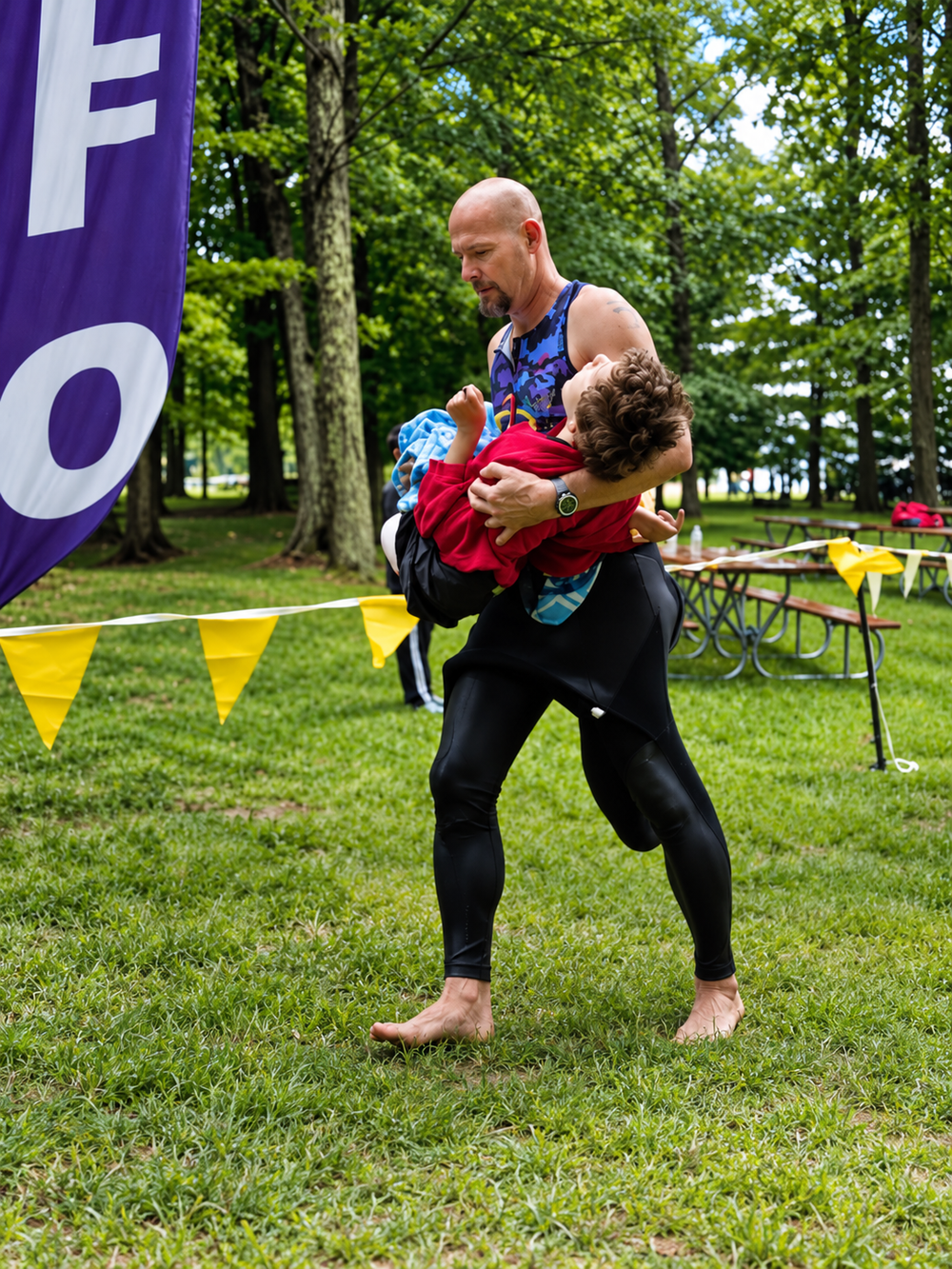 A barefoot man wearing a sleeveless top and wetsuit leggings carries a child in his arms across a grassy park.