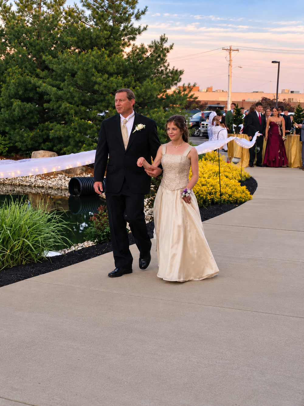 A man in a black tuxedo and a girl in a formal cream gown walk arm-in-arm along a concrete path, with other formally dressed people visible in the background.