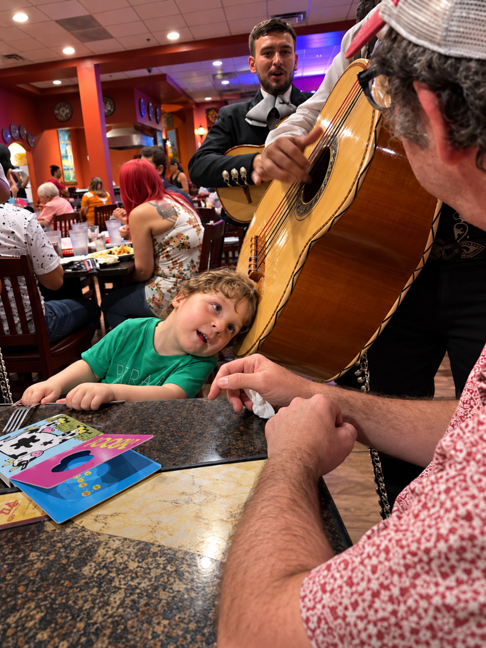 A small child with their head resting against a musician’s large acoustic guitar in a restaurant, while a man in a patterned shirt sits at the table in the foreground.