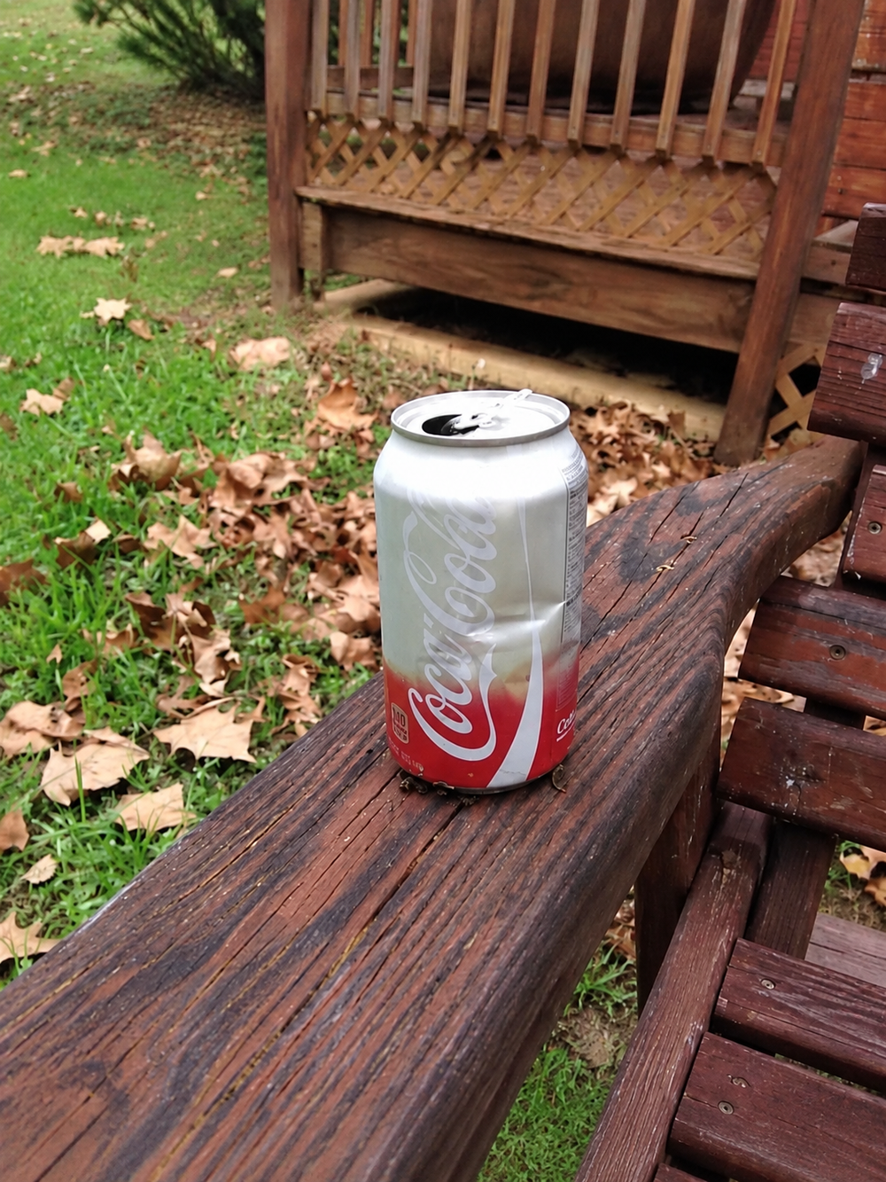 A soda can rests on an outdoor wooden railing marked by a dark circular wear ring.