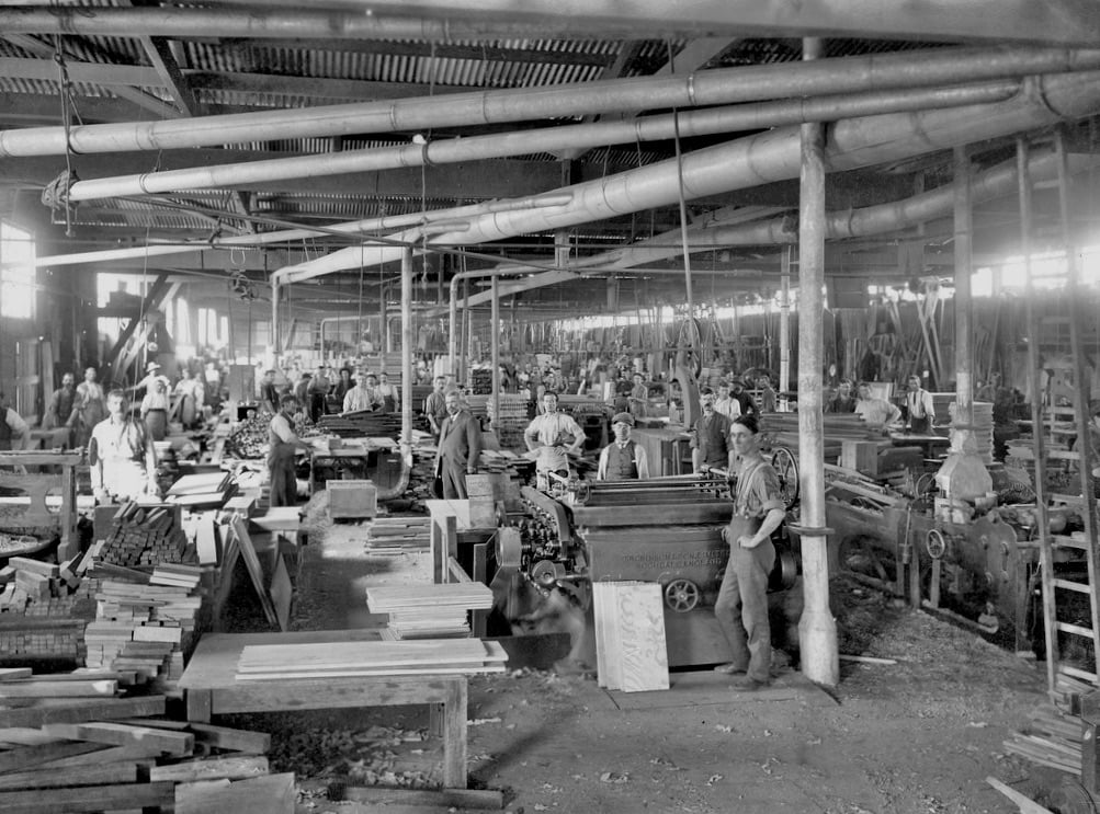 Workers in a wood manufacturing factory operate industrial machinery and handle stacks of lumber.