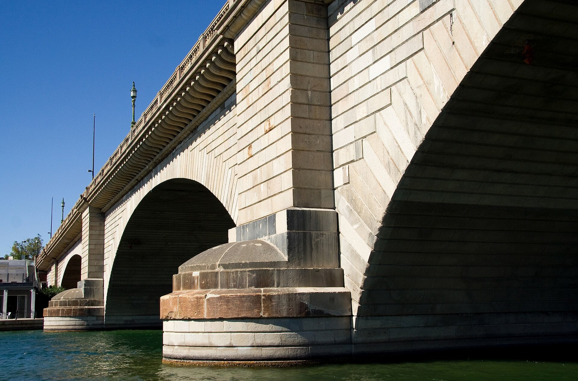 A low-angle view of a stone arch bridge spanning a river under a clear blue sky.