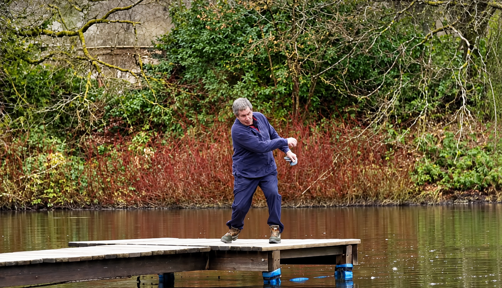 A man in a blue jacket and pants stands on a wooden dock and tosses a blue disc into the water.