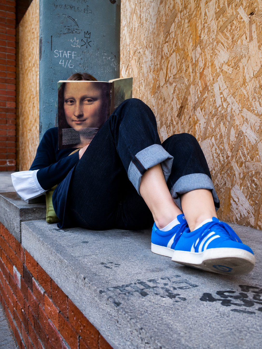 A person sitting on a ledge with their face obscured by a book featuring an image of the Mona Lisa.