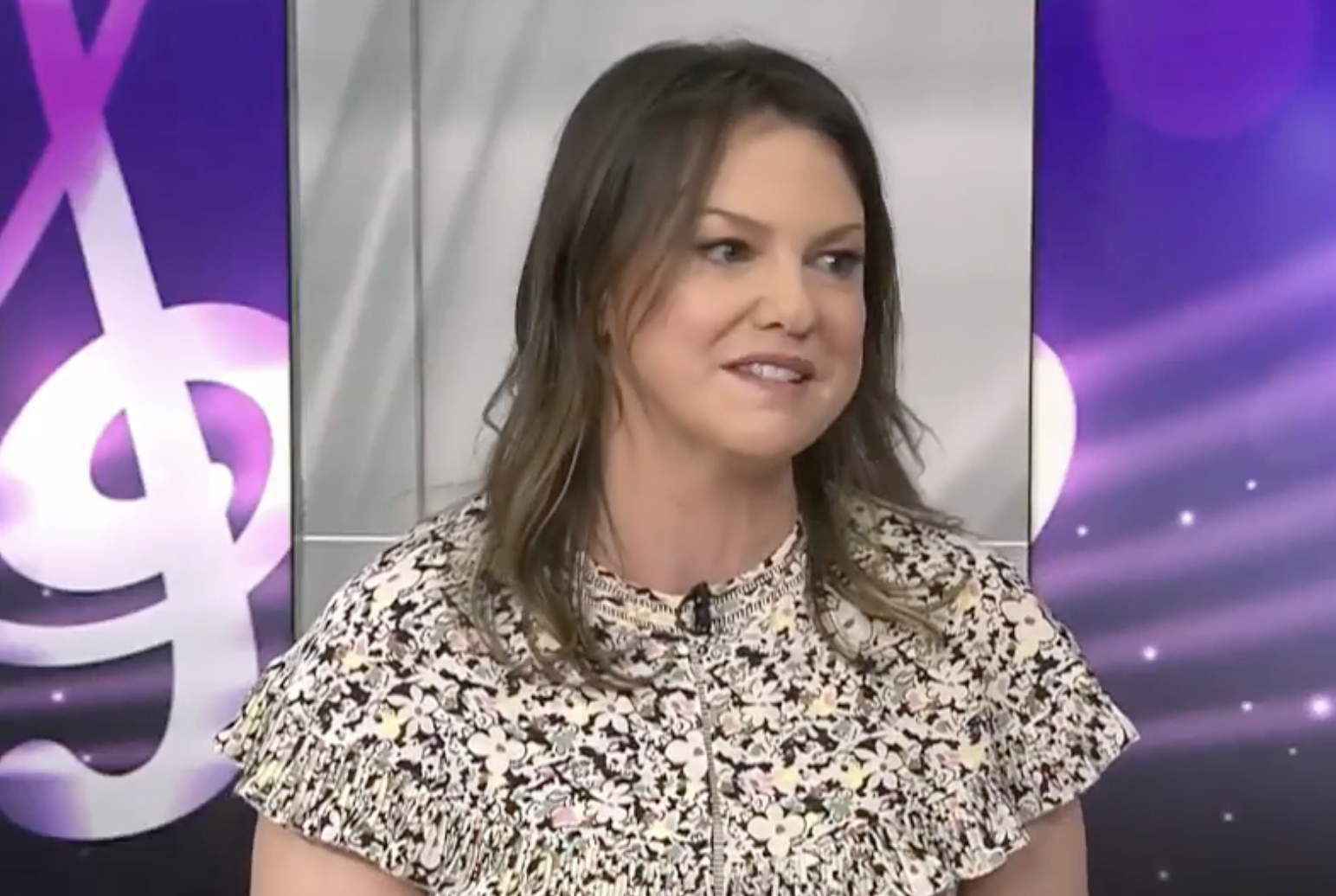 A woman with shoulder-length brown hair wearing a floral print top speaks in front of a purple backdrop with musical note graphics.
