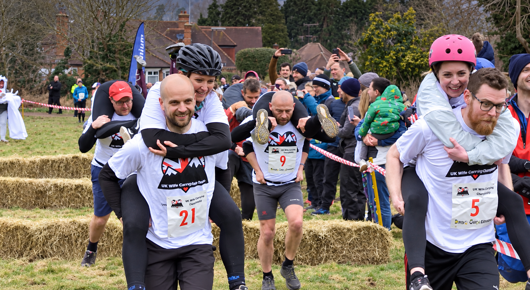 Three men participating in a wife-carrying race, each carrying a woman on their back, running past spectators on a grass field with hay bales.