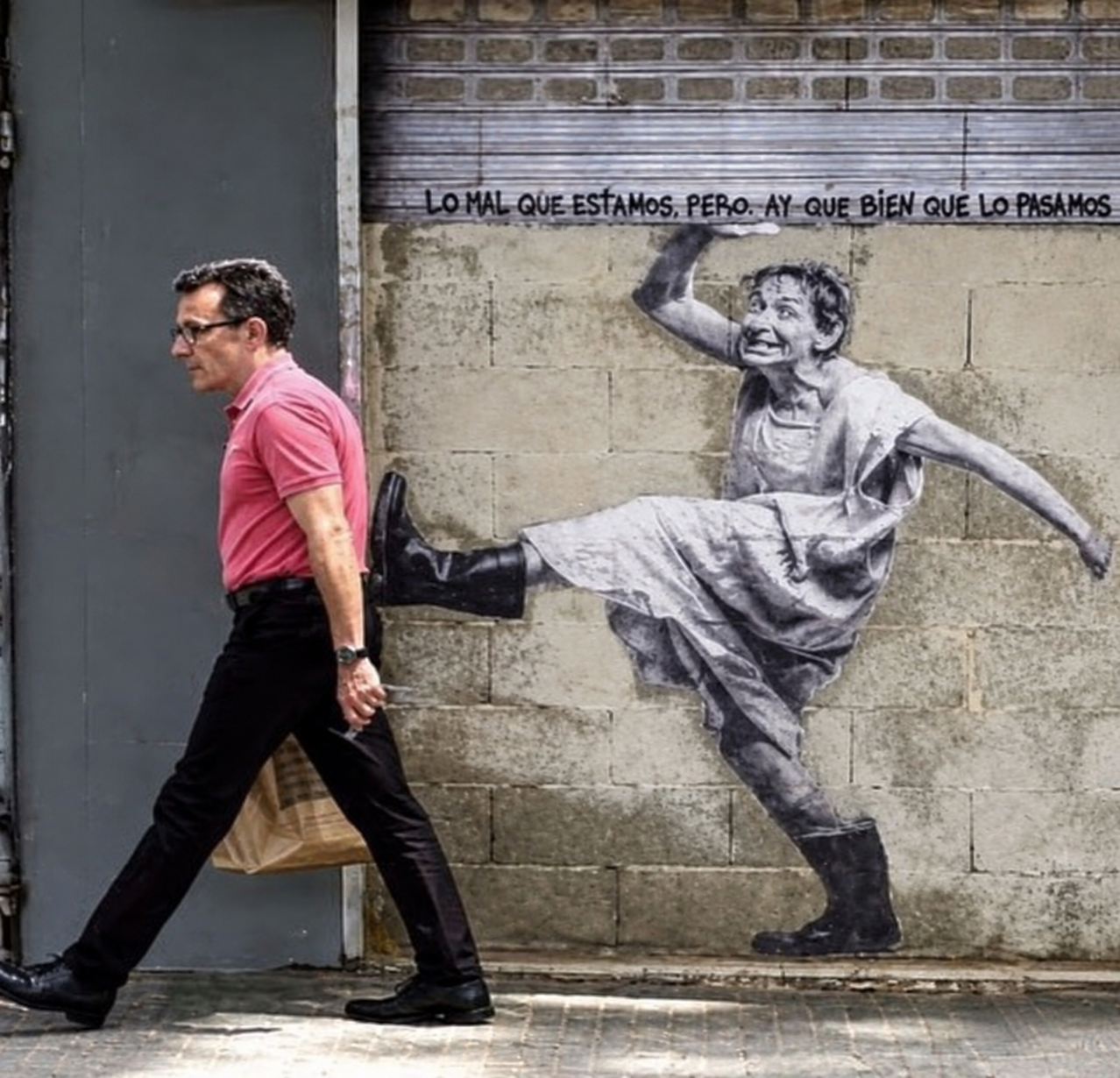 A man walking past a wall mural of a person in a dress and boots appearing to kick him, with the Spanish text “LO MAL QUE ESTAMOS, PERO, AY QUE BIEN QUE LO PASAMOS” written above.