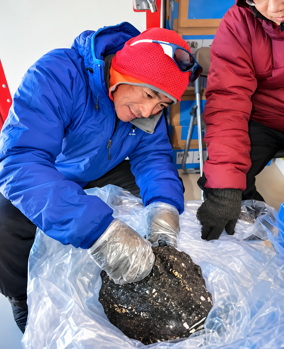 A person wearing a blue jacket and red beanie with plastic gloves examines a large, dark, textured rock inside a plastic bag.