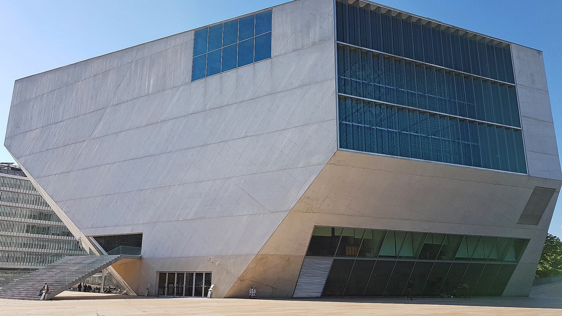 A large, angular modern building with concrete walls and glass facade sections under a clear blue sky.