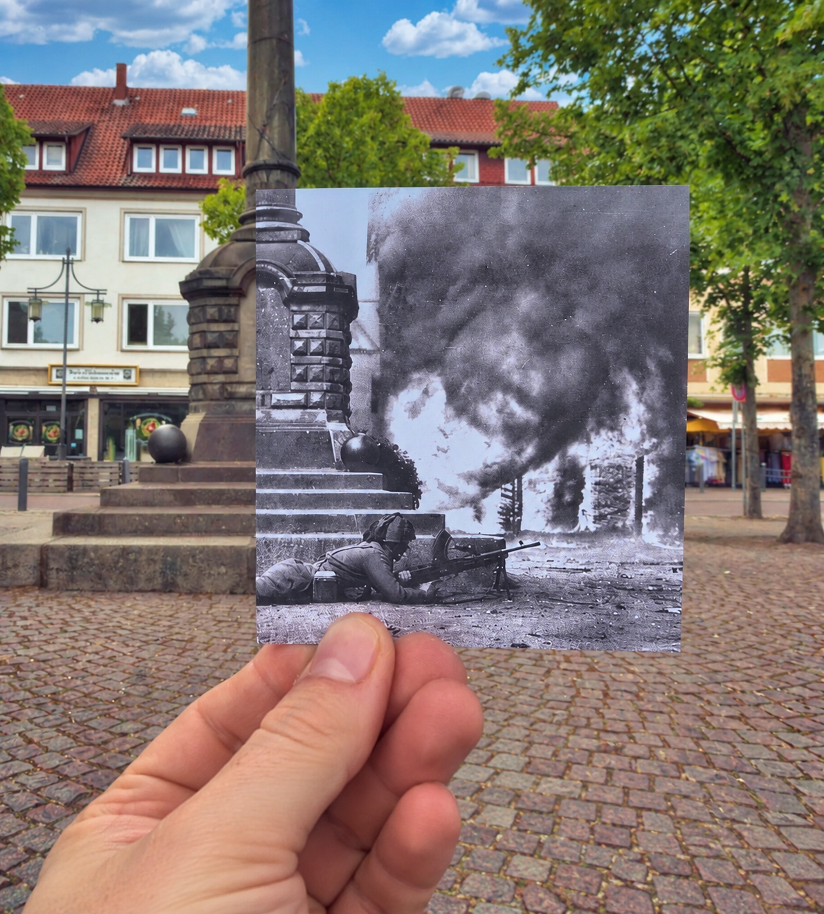 A hand holds a historical black-and-white photograph of a soldier prone with a machine gun in front of a burning building, perfectly aligned with the real-world stone monument in the background.