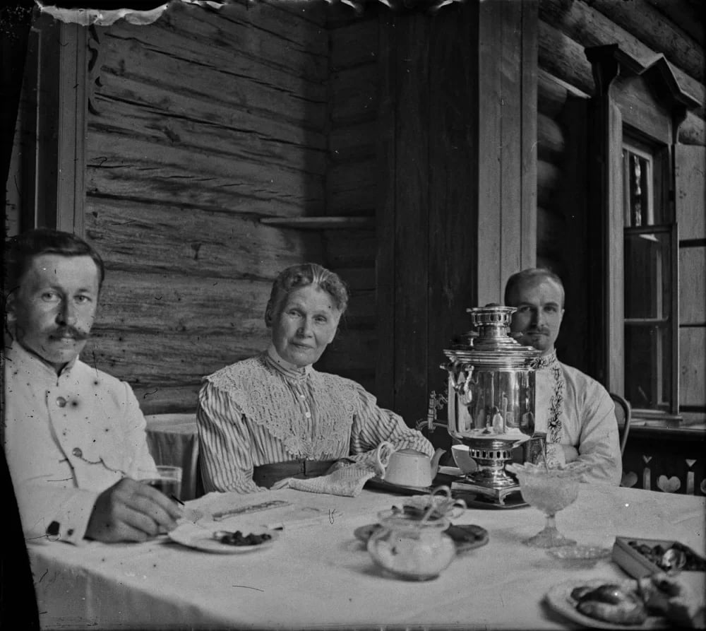 Three people sitting at a table set for tea with a samovar on a log cabin porch.