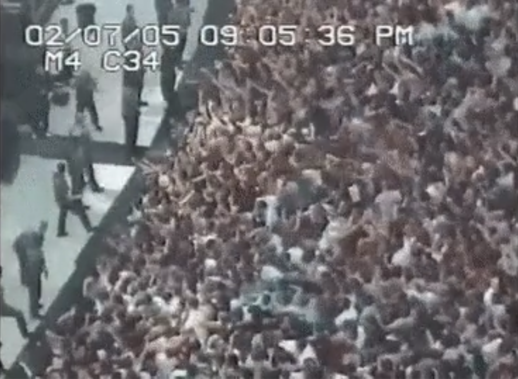 Security personnel stand near a dense, cheering crowd at a stadium event, captured in a black-and-white security camera view with a timestamp.